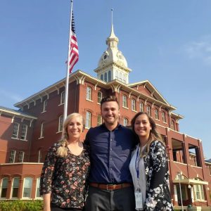 Three people smiling and standing outside in front of U.S. Flag pole and brick building.